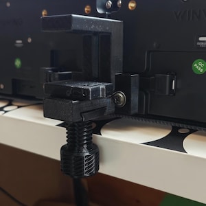 May include: A black, 3D-printed clamp with a threaded adjustment screw. The clamp is attached to a black electronic device. The device has a green QC Pass sticker.