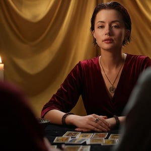 May include: A woman in a burgundy velvet top sits at a table with tarot cards, facing two people. A lit candle and gold drapery are in the background. She wears a necklace and bracelets. The scene suggests a fortune-telling or spiritual reading.