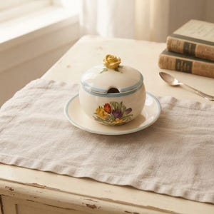 Könnte beinhalten: Eine cremefarbene Zuckerdose aus Keramik mit passender Untertasse und Deckel. Der Deckel ist mit einer skulptierten gelben Rose verziert. Die Dose und die Untertasse zeigen ein buntes Blumenmuster aus Tulpen und Narzissen sowie einen hellblauen Rand.