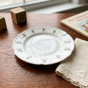 May include: A white ceramic plate with a scalloped edge and a repeating pattern of small, colorful illustrations of anthropomorphic animals dressed in clothing around the rim. The center of the plate is plain white and smooth.