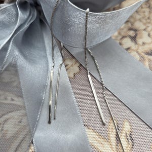May include: Silver threader earrings with a linear bar design. The earrings hang from delicate chains. They are displayed against a backdrop of a grey ribbon and floral patterned fabric.