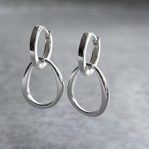 May include: A pair of silver-toned hoop earrings, each featuring a small circular hinged hoop at the top connected to a larger, slightly irregular, open teardrop-shaped pendant. The metal has a smooth, polished, and reflective finish throughout.
