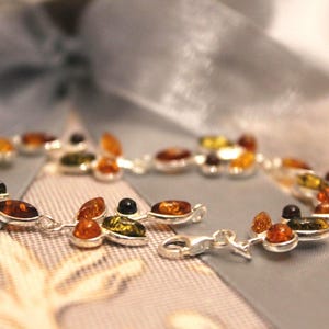 May include: A silver bracelet featuring a series of amber-coloured stones. The stones vary in colour, including shades of yellow, orange, and brown. The bracelet is set against a soft, blurred background.