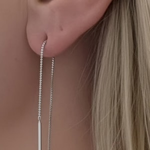 May include: A silver threader earring featuring a delicate chain that passes through the earlobe, ending in a slender, polished vertical bar. The earring is crafted from a bright, reflective silver-toned metal, creating a clean and linear aesthetic.
