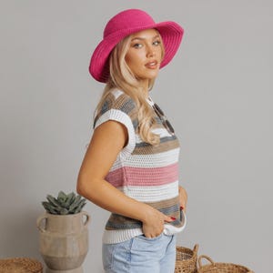 May include: A woman wearing a fuchsia sun hat and a striped, short-sleeved sweater. The sweater features horizontal stripes in white, pink, brown, and grey. Sunglasses are tucked into the neckline. The woman is wearing blue jeans.