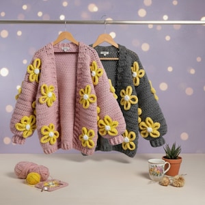 Op de afbeelding: Twee openvallende vesten, één roze en één grijs, versierd met grote gele en witte bloemenapplicaties. De truien hangen aan houten kleerhangers tegen een zachte paarse achtergrond. Een bol roze garen, een gele bol garen en een klein gehaakt vierkant staan op de voorgrond.