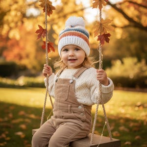 Könnte beinhalten: Ein Kind trägt eine weiße Strickmütze mit orangefarbenen und blauen Streifen und einem weißen Bommel, einen cremefarbenen Pullover und eine braune Cord-Latzhose. Das Kind sitzt auf einer Holzschaukel, mit Herbstblättern und einem verschwommenen Hintergrund.