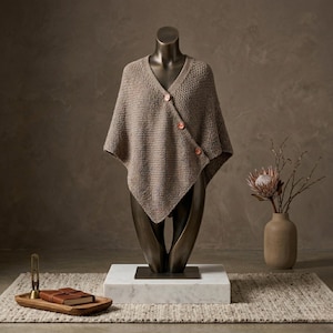 May include: A neutral-toned, knitted poncho with a V-neck and three wooden buttons. The poncho is displayed on a bronze mannequin, set against a neutral background. A vase with dried flowers and a wooden tray with a leather-bound book are also visible.
