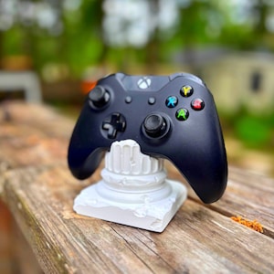 May include: A black Xbox controller rests on a white, sculpted stand. The controller features colored buttons and directional controls. The stand has a classical column design, and the background is a blurred outdoor scene.