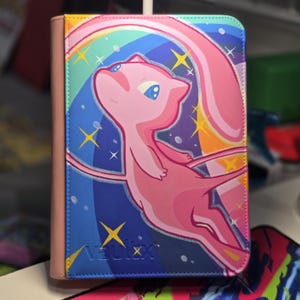 Può includere: Un raccoglitore rettangolare per carte collezionabili con dorso rosa e una copertina che raffigura un personaggio Mew rosa che fluttua su uno sfondo arcobaleno vibrante e un cielo blu scuro stellato. La parola vaulx è impressa in basso a sinistra.