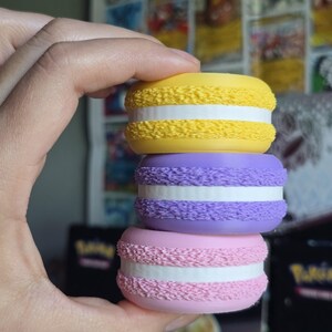 May include: A hand holds a stack of three macarons. The top macaron is yellow, the middle one is purple, and the bottom one is pink. Each macaron has a white filling and a textured, ruffled edge. The macarons are stacked on top of each other.