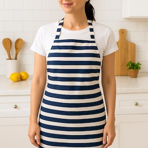 May include: A navy blue and white striped apron. The apron has a bib top and a waist tie. The stripes are horizontal and evenly spaced. The apron is worn over a white t-shirt. The background includes a white countertop, wooden utensils, and a cutting board.