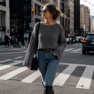 May include: A woman wearing a gray, open-weave sweater, blue jeans, and black boots, crossing a city street. She is wearing sunglasses and carrying a black handbag. The sweater has long sleeves and a cropped fit. The jeans are a light wash.