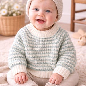 Könnte beinhalten: Ein lächelndes Baby trägt einen hellblau-weiß gestreiften Pullover, eine beige Mütze und eine beige Hose. Das Baby sitzt auf einem weichen, hellfarbigen Teppich. Der Pullover hat weiße Bündchen und einen weißen Kragen.