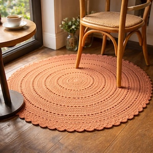 Pode incluir: Um tapete redondo de crochê cor de pêssego com borda ondulada. O tapete apresenta círculos concêntricos de pontos de crochê, criando uma superfície texturizada. O tapete está colocado sobre um chão de madeira, com uma cadeira e uma mesa ao fundo.