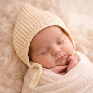 May include: A sleeping baby wearing a cream-coloured knitted bonnet and wrapped in a matching blanket. The baby's eyes are closed, and the hands are gently resting near the face. The background is a soft, textured surface.