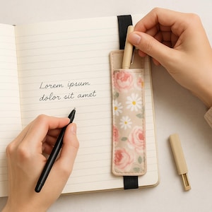 Könnte beinhalten: Ein offenes Notizbuch mit linierten Seiten und dem Text "Lorem ipsum dolor sit amet". Ein floraler Stifthalter mit rosa Rosen und weißen Gänseblümchen ist angebracht. Ein schwarzer Stift wird verwendet, und ein beiger Stift wird aus dem Halter entfernt.