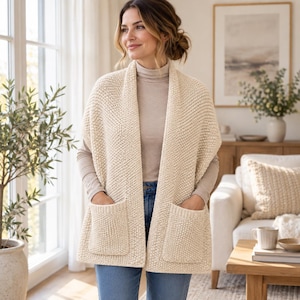 Può includere: Un gilet in maglia color crema senza maniche, aperto sul davanti, caratterizzato da un motivo a maglia testurizzato e due grandi tasche applicate quadrate. Il capo è indossato sopra una maglia a collo alto beige a maniche lunghe e jeans blu.