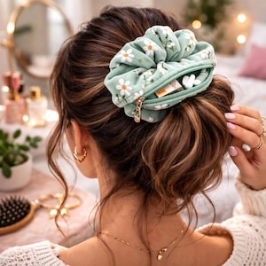 Peut inclure: Un chouchou vert menthe avec un motif de marguerites blanches et une poche zippée. Le chouchou est porté en chignon. La fermeture éclair est dorée. Le chouchou est un accessoire pour cheveux.