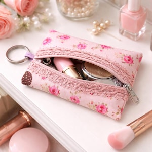 May include: Pink floral print cosmetic bag with a zipper, lace trim, and a key ring. The bag is open, revealing lipstick and a compact. A makeup brush and nail polish are in the background.