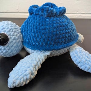 May include: A handmade plush turtle in shades of blue. The turtle has a light blue head and limbs, a dark blue shell, and large black eyes. The soft, textured material gives it a cuddly appearance.