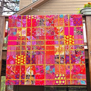 Op de afbeelding: Een levendige, handgemaakte quilt met een patchwork-ontwerp. De quilt heeft een raster van kleurrijke vierkanten in tinten rood, roze, oranje en paars, met goudkleurige stiksels. De vierkanten tonen bloemen- en patroonstoffen.
