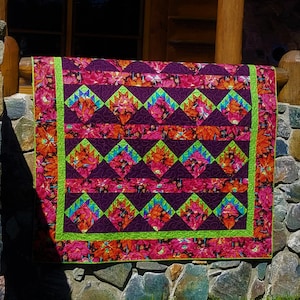 May include: A colorful quilt with a floral pattern in shades of pink, orange, and purple. The quilt features a geometric design with diamond shapes and a bright green border. The quilt is displayed outdoors, draped over a stone wall.