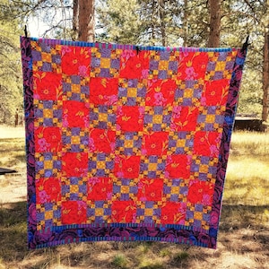 May include: A red, yellow, and purple quilt with a floral pattern. The quilt is hanging on a clothesline in a wooded area.