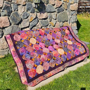 May include: A colorful quilt with a geometric pattern in shades of purple, pink, and orange. The quilt is made of fabric scraps and is displayed on a wooden bench.