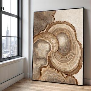 May include: A framed abstract art print featuring a close-up view of tree rings in shades of brown and beige. The artwork has a natural, organic aesthetic, with detailed textures and patterns. The print is displayed in a room with a window.