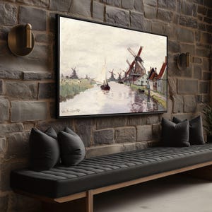 Könnte beinhalten: Gerahmtes Landschaftsgemälde mit Windmühlen an einem Kanal, mit einem kleinen Segelboot und Figuren. Das Kunstwerk befindet sich in einem schwarzen Rahmen, der an einer Backsteinmauer über einer schwarzen Bank mit grauen Kissen angebracht ist.