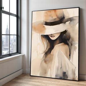 Puede incluir: Pintura a la acuarela enmarcada de una mujer con sombrero de ala ancha. La obra de arte utiliza una paleta de tonos beige, marrón y blanco. El rostro de la mujer está parcialmente oculto por el sombrero. La pintura se exhibe en una habitación con una ventana con vistas a un paisaje urbano.