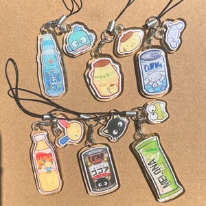 May include: A collection of clear acrylic charms featuring various cartoon food and drink designs. The charms include a blue soda bottle, a fish, a pudding cup, a can of soda, a bottle of juice, a black cat, a popsicle, and a green ice cream bar.