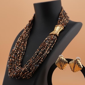 May include: A multi-strand necklace featuring small brown, black, and metallic beads, secured by a gold-toned ribbed metal clasp. The set includes a pair of matching gold-toned ribbed clip-on earrings with a textured, fan-like design.