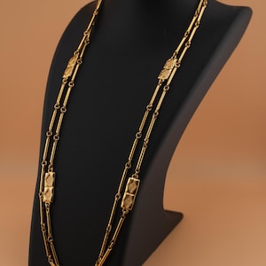 May include: A long, gold-colored necklace displayed on a black mannequin. The necklace features a repeating pattern of rectangular and cylindrical links. The rectangular links have a textured design. The necklace is a vintage or antique style.