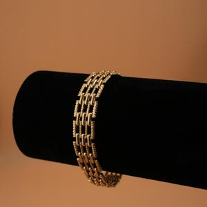 May include: A gold-toned bracelet with a rectangular link design. The bracelet is displayed on a black velvet surface against a warm brown background. The jewelry piece has a textured appearance.