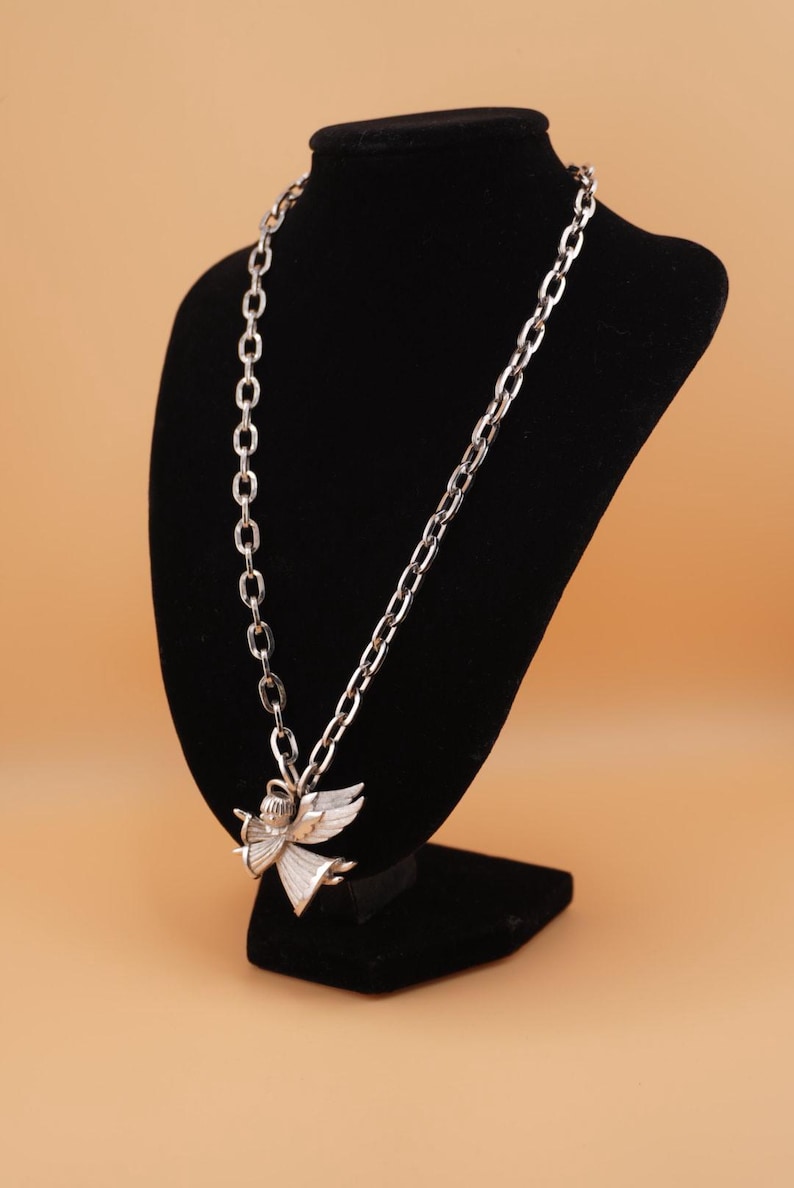 Op de afbeelding: Zilverkleurige ketting met een engel-hanger, tentoongesteld op een zwarte fluwelen buste. De ketting heeft twee verschillende stijlen, &eacute;&eacute;n met ovale schakels en de andere met een dubbele schakel.