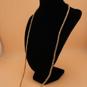 Puede incluir: Un collar dorado con un diseño de cuerda trenzada, exhibido en un busto de joyería negro. El collar tiene una apariencia delicada y texturizada y se encuentra sobre un fondo color melocotón.