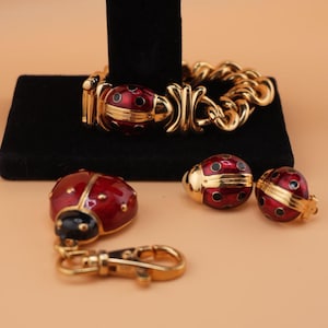 May include: A gold-coloured jewellery set featuring a bracelet, earrings, a keychain, and a pendant, all with a ladybug theme. The ladybug elements are red with black spots and accents. The bracelet is on a black stand.
