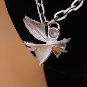 Op de afbeelding: Zilverkleurige ketting met een bloemvormige hanger. De hanger heeft een gestileerd bloemontwerp met meerdere bloemblaadjes en een centraal element. De ketting is een schakelontwerp. De hanger heeft het Trifari-merk.
