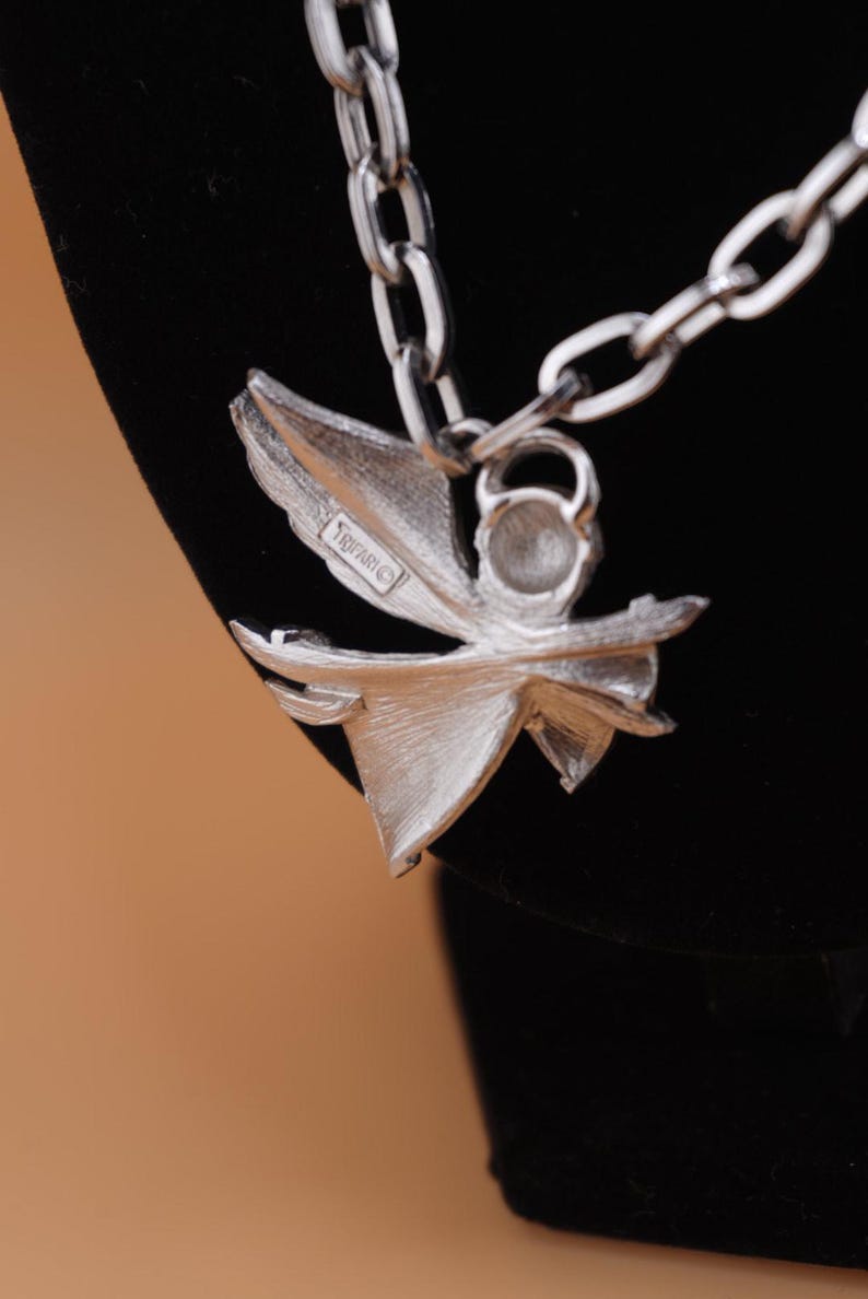 Op de afbeelding: Zilverkleurige ketting met een bloemvormige hanger. De hanger heeft een gestileerd bloemontwerp met meerdere bloemblaadjes en een centraal element. De ketting is een schakelontwerp. De hanger heeft het Trifari-merk.