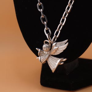 Op de afbeelding: Zilverkleurige engel hanger ketting. De hanger heeft een engel met uitgestrekte vleugels en een vloeiende jurk. De ketting heeft een schakelontwerp. De sieraden worden getoond op een zwart fluwelen oppervlak tegen een bruine achtergrond.