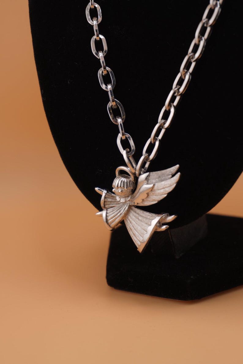 Op de afbeelding: Zilverkleurige engel hanger ketting. De hanger heeft een engel met uitgestrekte vleugels en een vloeiende jurk. De ketting heeft een schakelontwerp. De sieraden worden getoond op een zwart fluwelen oppervlak tegen een bruine achtergrond.