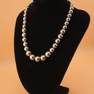 May include: A silver-colored beaded necklace displayed on a black velvet bust. The necklace features a graduated design with larger beads at the front, decreasing in size towards the clasp. The beads have a polished, reflective surface.