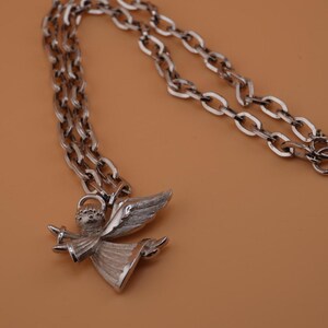 Op de afbeelding: Zilverkleurige ketting met een engel hanger. De engel heeft gedetailleerde vleugels en een vloeiende jurk. De ketting heeft een schakelontwerp. De achtergrond is effen bruin. Een elegant sieraad.