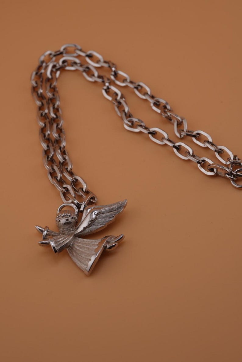 Op de afbeelding: Zilverkleurige ketting met een engel hanger. De engel heeft gedetailleerde vleugels en een vloeiende jurk. De ketting heeft een schakelontwerp. De achtergrond is effen bruin. Een elegant sieraad.
