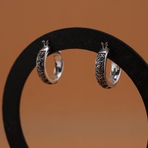 May include: Two silver hoop earrings with an intricate, engraved pattern. The earrings are displayed against a warm brown background, showcasing their detailed design and polished finish. The earrings are a classic jewelry item.