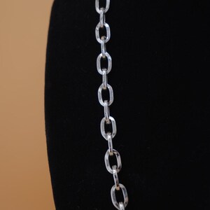 Op de afbeelding: Een zilverkleurige ketting met grote ovale schakels. De ketting wordt gepresenteerd tegen een zwarte achtergrond, waardoor het reflecterende oppervlak wordt benadrukt. De ketting is een eenvoudig maar elegant sieraad.