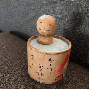 May include: A wooden Japanese Kokeshi doll with a painted face and blue hair sits atop a cylindrical container. The container has Japanese calligraphy and a painted design in red, orange, and green. The doll is a traditional Japanese craft.