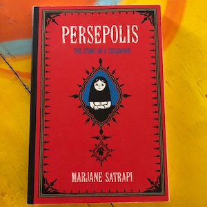 May include: A red book with a black border and the title "Persepolis: The Story of a Childhood" in white text. The cover features a black and white illustration of a girl wearing a headscarf. The author's name, "Marjane Satrapi", is printed in white text at the bottom of the cover.